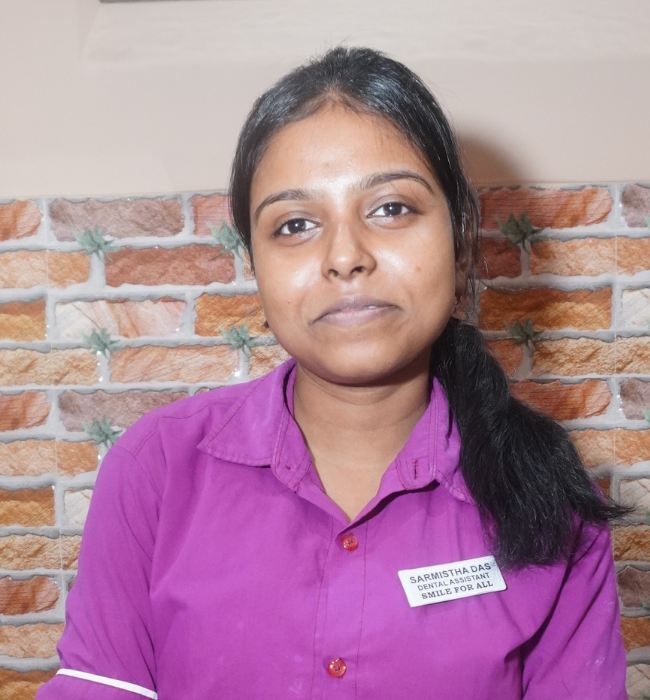 Female dental assistant in a purple uniform smiling softly against a brick-patterned clinic wall.