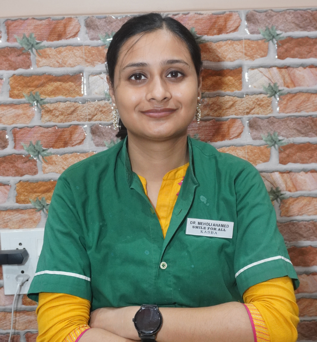 Female dental professional in a green uniform posing confidently with folded arms against a brick-patterned clinic wall.
