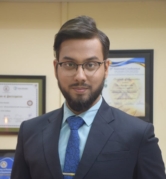 Male dental professional in a formal suit posing confidently in front of framed certificates in a clinic office.