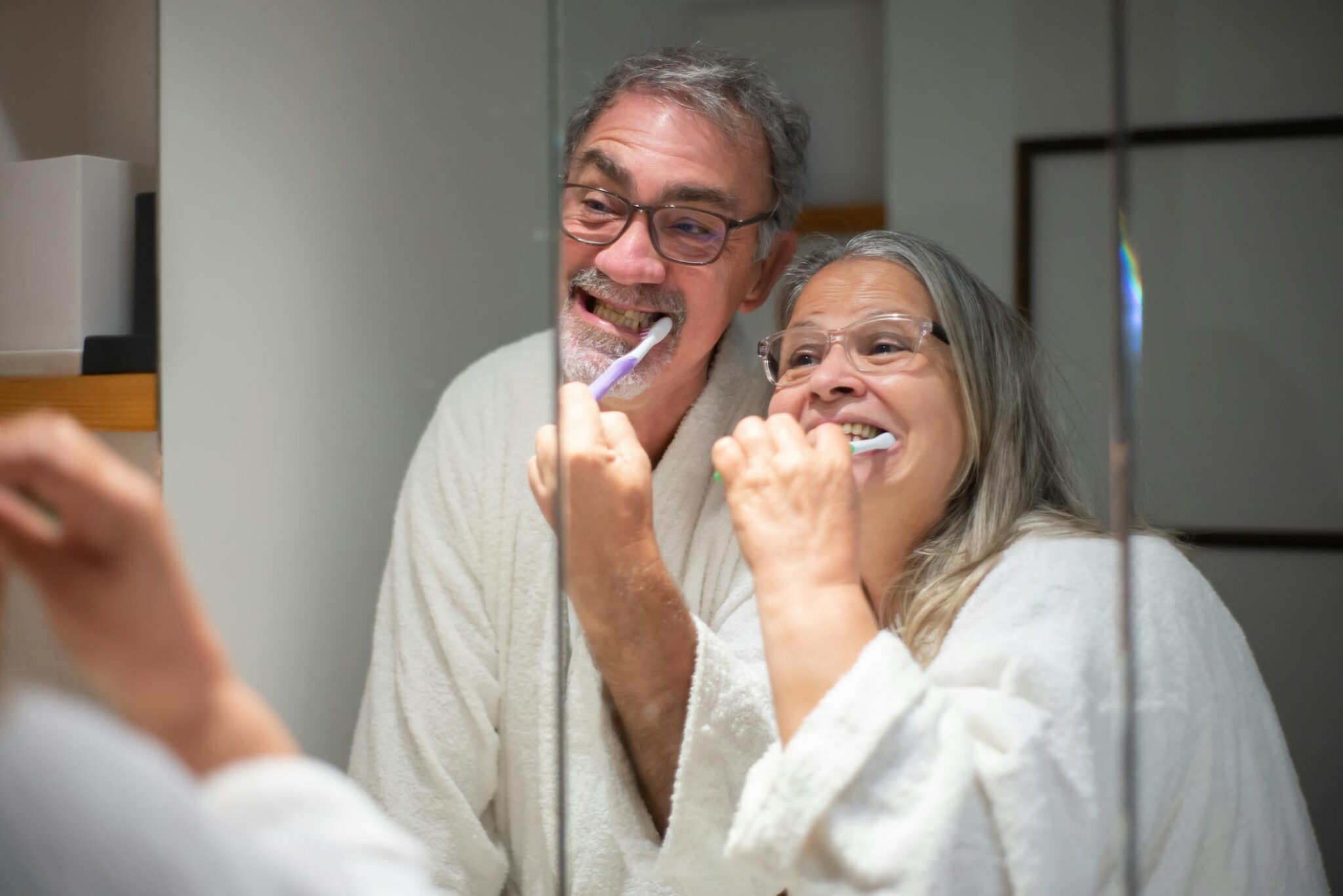 Senior couple smiling while brushing their teeth together in front of a bathroom mirror.