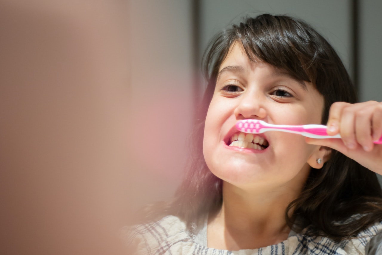 Young child brushing teeth with a pink toothbrush while smiling in front of the bathroom mirror.