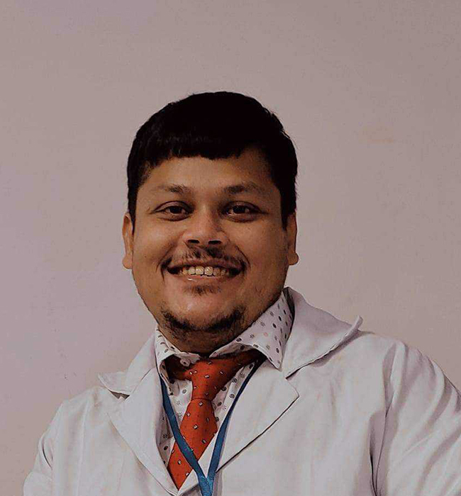 Male dental professional in a white coat smiling warmly while posing against a plain clinic background.