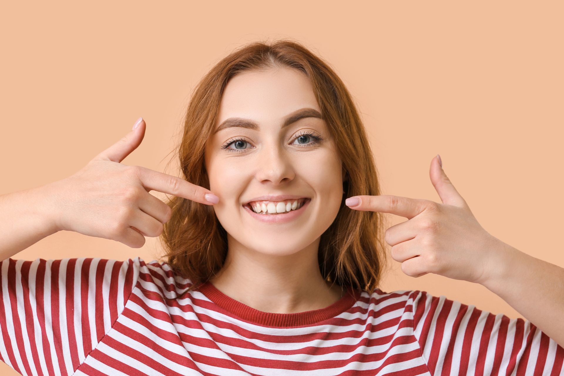 Young woman smiling and pointing to her teeth, showing a healthy and confident smile against a peach background.