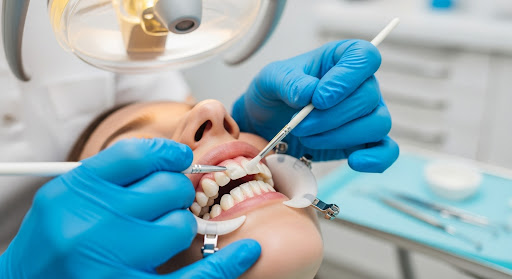 Dentist performing a dental procedure on a patient using instruments and a cheek retractor under clinic lighting.