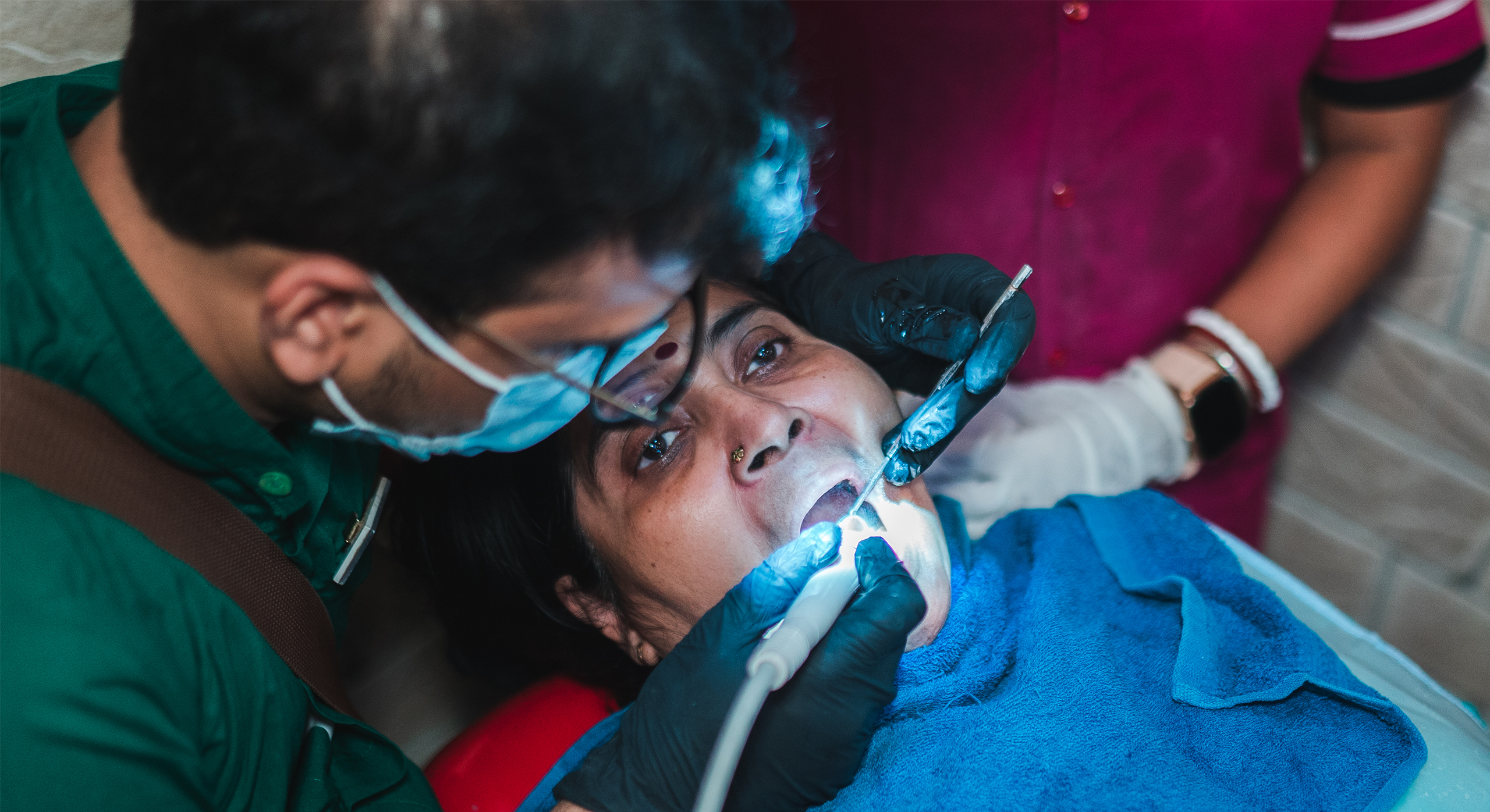 Dentist performing a detailed oral procedure on a patient with bright examination light and support staff nearby.